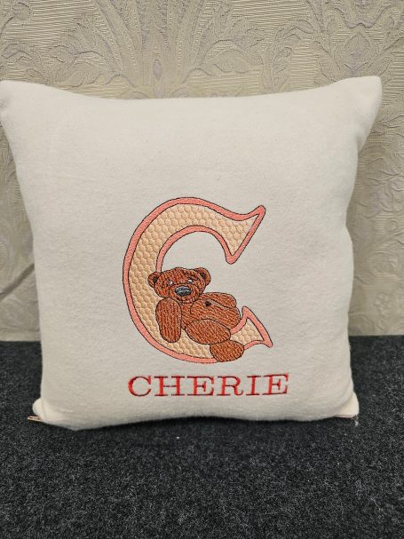 Light-coloured cushion with an embroidered letter "C" and a teddy bear design, labelled "CHERIE".
