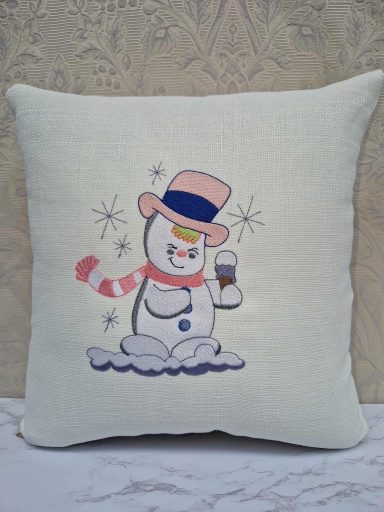 A decorative cushion featuring a snowman holding a drink, wearing a pink hat and scarf.