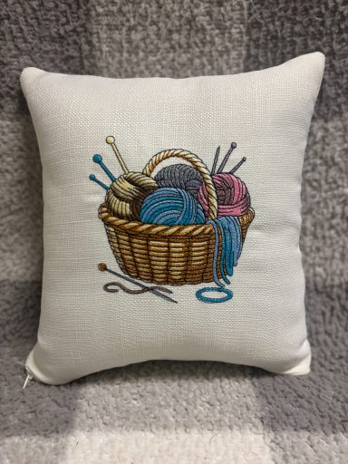 Embroidered cushion featuring a basket of yarn and knitting needles.