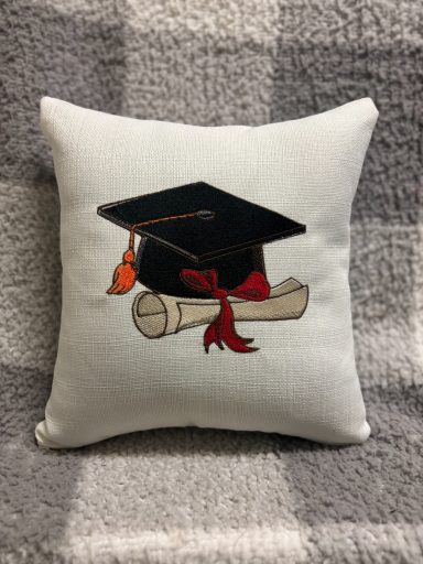 Embroidered cushion featuring a graduation cap and diploma with a red ribbon.