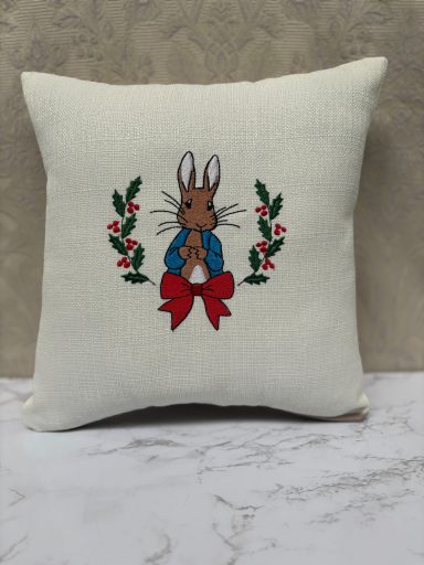 A decorative cushion featuring an embroidered rabbit in a blue jacket surrounded by foliage.