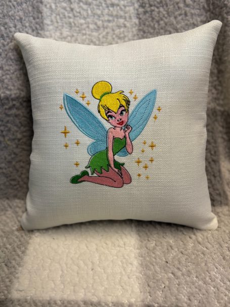 A decorative cushion featuring Tinker Bell with turquoise wings and a mischievous pose.