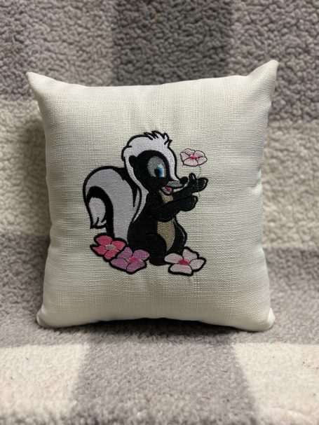 Cartoon skunk on a cushion, wearing pink slippers and holding a small object.