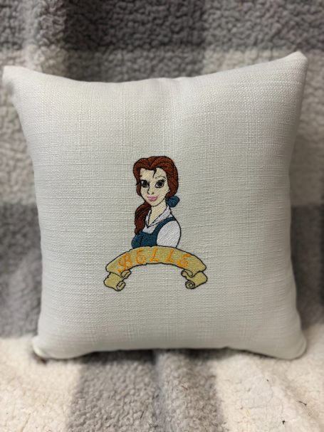 White cushion featuring an embroidered character with brown hair and a blue dress.