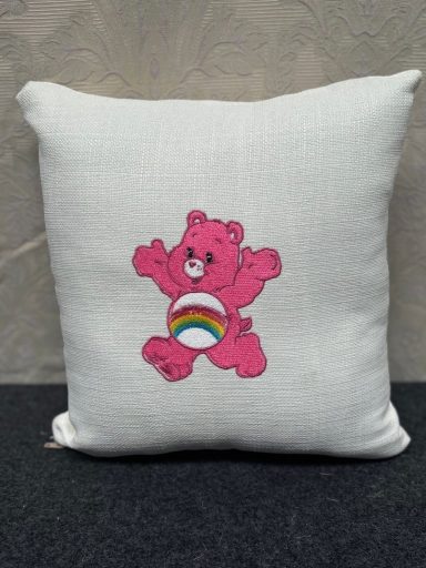 A soft cushion featuring a pink bear with a rainbow on its belly, smiling and jumping.