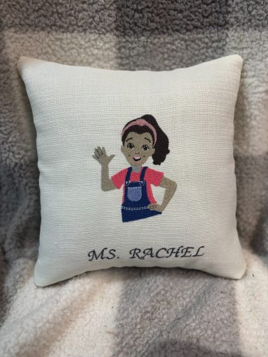 A decorative cushion featuring a cartoon character named Ms. Rachel waving.