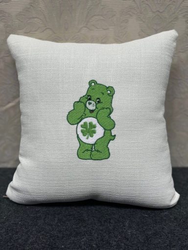 A white cushion featuring a green bear with a four-leaf clover on its belly.