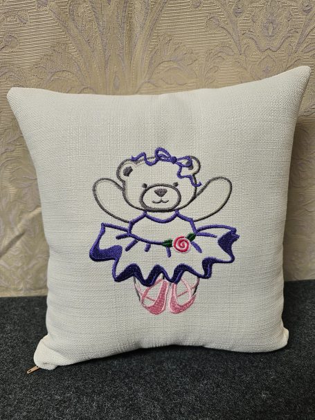 White cushion with a cartoon bear wearing a blue tutu and ballet shoes, holding its arms out.