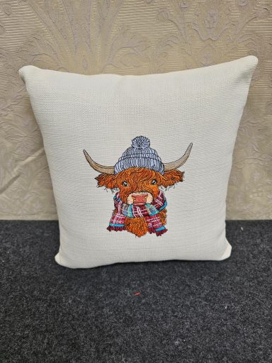 A white cushion with an embroidered Highland cow wearing a blue hat and scarf.