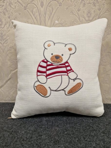 A white cushion featuring a teddy bear in a red and white striped sweater.