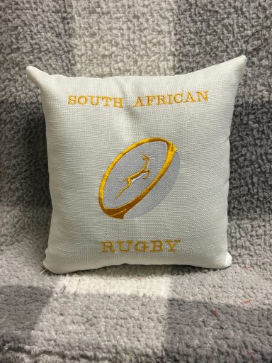 White cushion with yellow embroidery reading "South African Rugby" and a rugby ball design.
