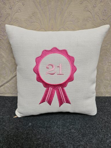 A cream cushion with a pink ribbon design featuring the number 21.