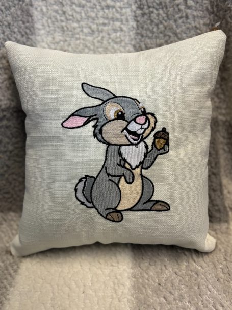 Grey rabbit cartoon character holding a snack, sitting on a decorative pillow.