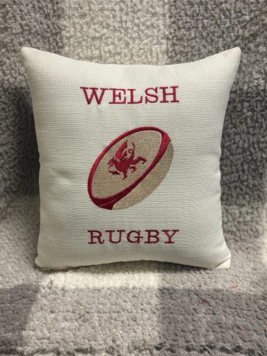 Cream cushion featuring red embroidered text "WELSH RUGBY" and a rugby ball design.