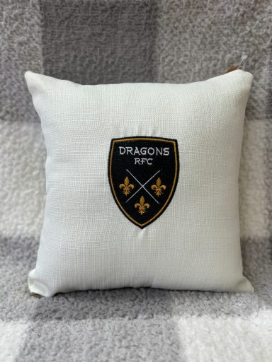 White cushion featuring an embroidered shield logo with black and yellow details.