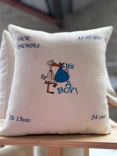 A decorative cushion featuring a stork and the text "It's a boy!" with birth details.