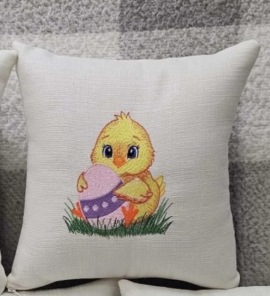Yellow chick holding a pink egg, sitting on green grass, on a decorative cushion.