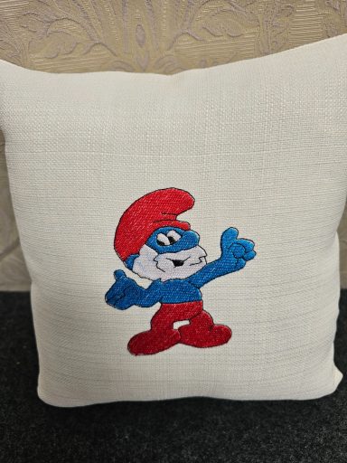 A beige cushion with an embroidered blue Smurf character in a red hat.