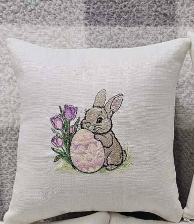A decorative cushion featuring a bunny beside a decorated egg and purple tulips.