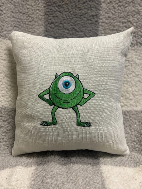 Green cartoon character with one eye and arms, standing confidently on a cushion.