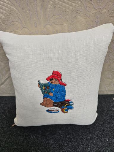 A decorative cushion featuring a bear in a red hat reading a book.
