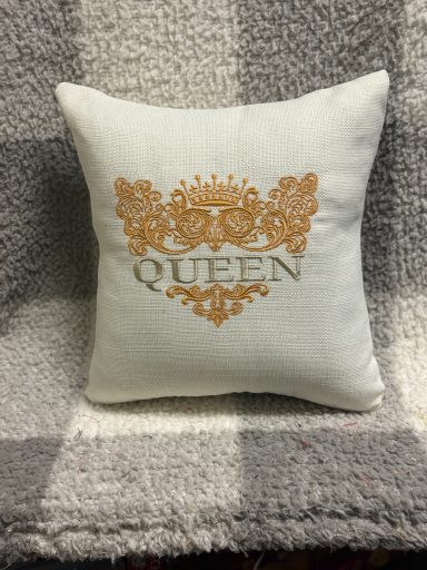White cushion with gold decorative pattern and the word "QUEEN" embroidered.