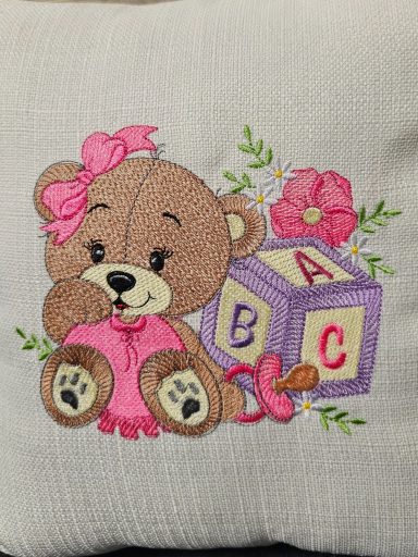 A cute bear sitting next to a colourful alphabet block and flowers, wearing a pink bow.