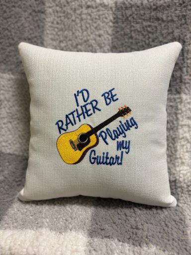 White cushion with blue text saying "I'd rather be playing my guitar!" and a yellow guitar illustration.