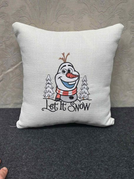 White cushion featuring a cartoon snowman with the text "Let It Snow" and pine trees.