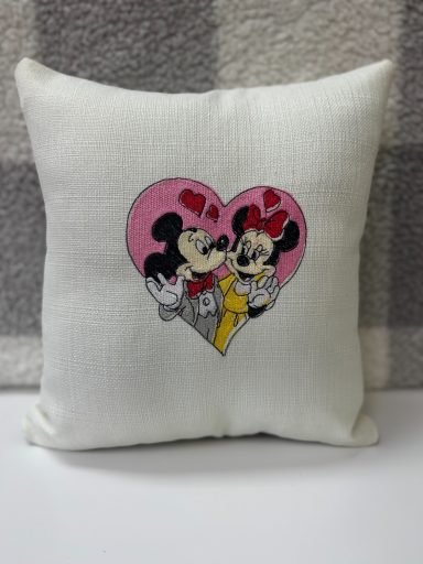 White cushion featuring Mickey and Minnie Mouse in a heart with pink accents.